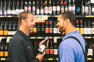 Operator holding bottle of wine talking to customer in front of wine bottle shelves