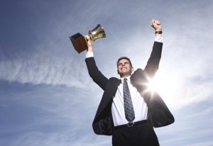 Man with hands up holding trophy, background is sky with sun shining